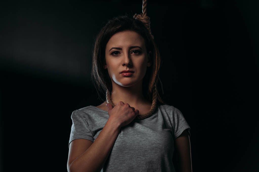frustrated woman looking at camera while standing with noose on neck on black background - Photo, Image
