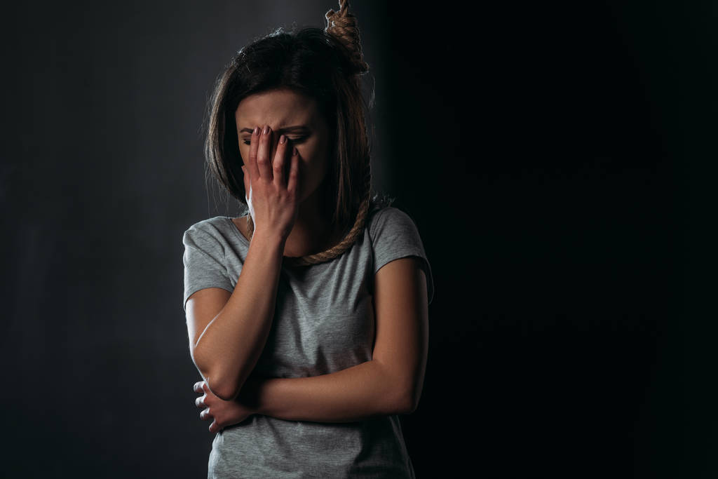 crying woman covering face with hand while standing with noose on neck on black background - Photo, Image
