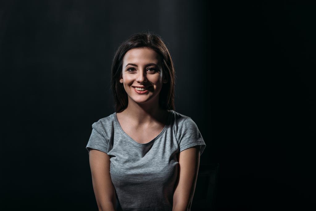 Ευτυχισμένη νεαρή γυναίκα χαμογελά στην κάμερα ενώ κάθεται στο σκοτάδι απομονωμένη στο μαύρο - Φωτογραφία, εικόνα