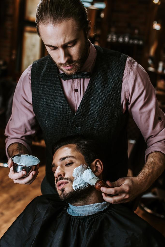 schöner Friseur, der Rasierschaum auf das Gesicht eines bärtigen Mannes aufträgt  - Foto, Bild