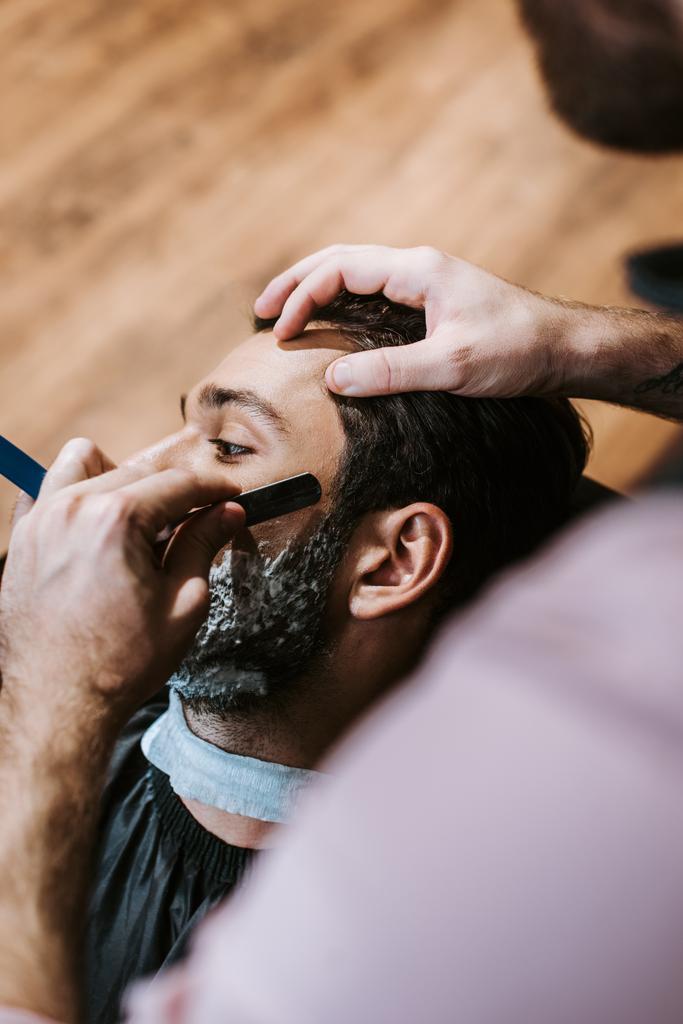 Selektiver Fokus des Barbiers rasiert Mann mit Rasierschaum im Gesicht  - Foto, Bild