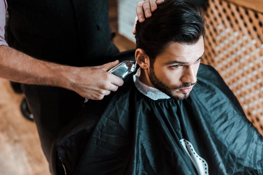barber holding trimmer while styling hair of handsome man in barbershop  - Photo, Image