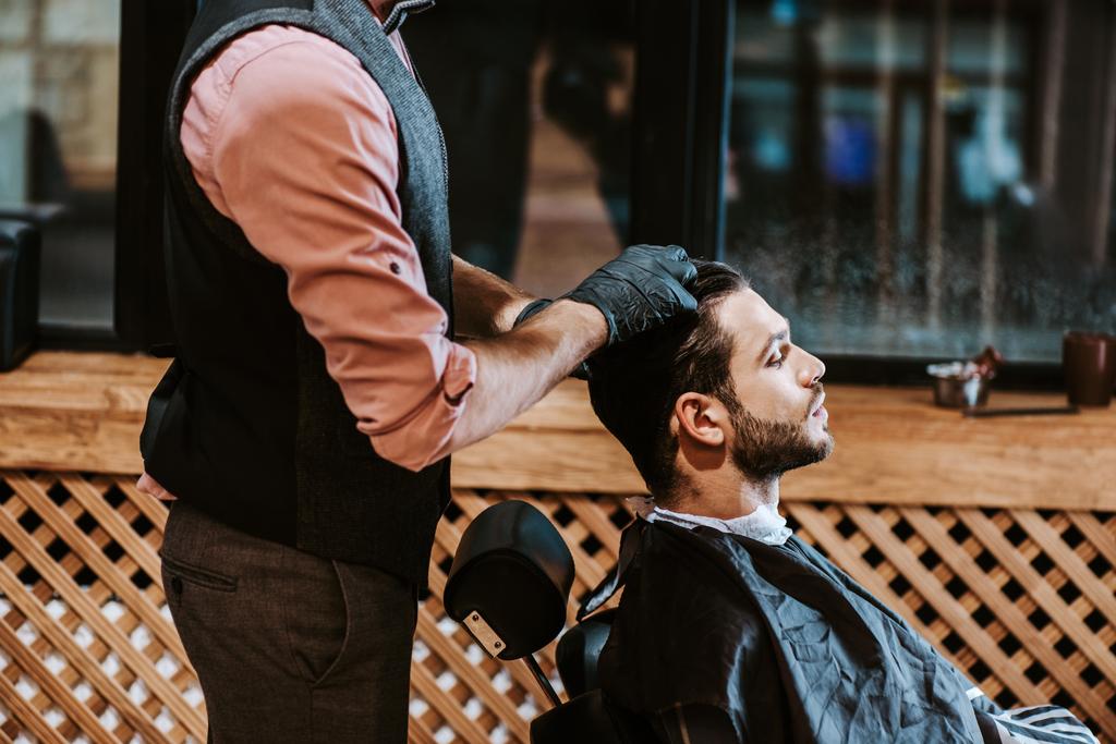 widok z boku brodaty mężczyzna siedzi obok fryzjera w czarnych rękawiczkach lateksowych  - Zdjęcie, obraz