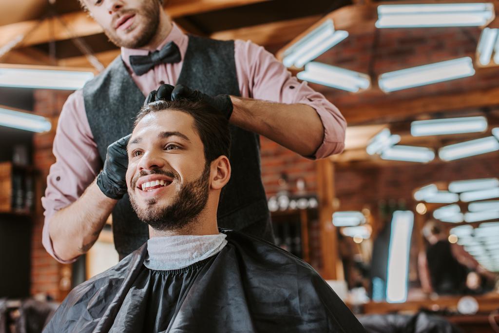 barber in black latex gloves touching hair of happy man in barbershop  - Photo, Image