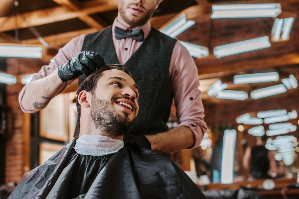 brodaty fryzjer w czarnych rękawiczkach lateksowych dotykający włosów szczęśliwego mężczyzny w salonie fryzjerskim  - Zdjęcie, obraz