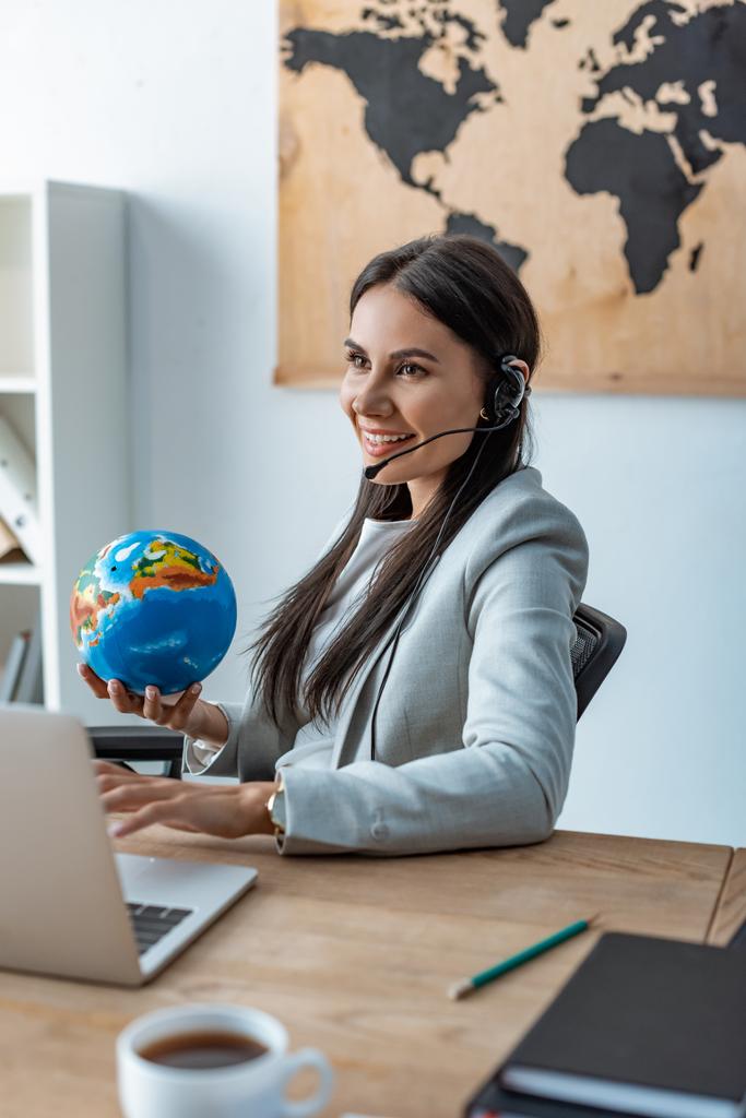 smiling travel agent holding globe while sitting at workplace - Foto, Bild