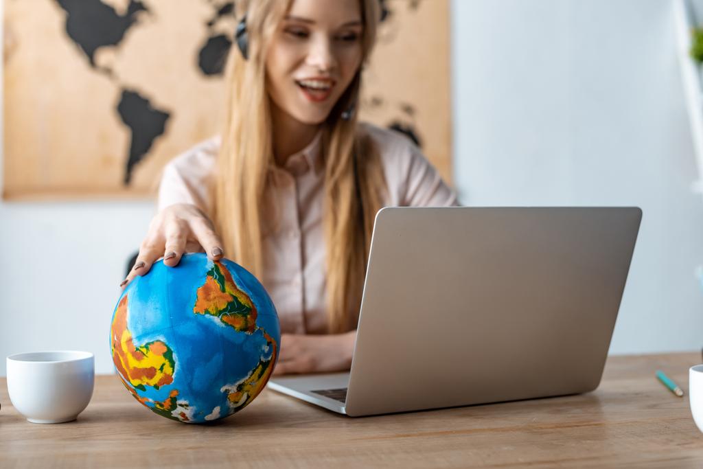 selective focus of smiling travel agent touching globe while looking at laptop - Photo, Image