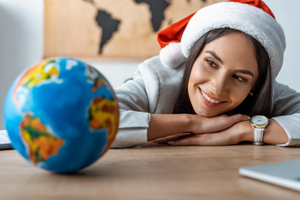 smiling travel agent looking at globe while sitting at workplace - Photo, Image