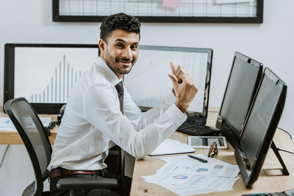 smiling bi-racial trader showing thumbs up and sitting near computers with graphs  - Photo, Image