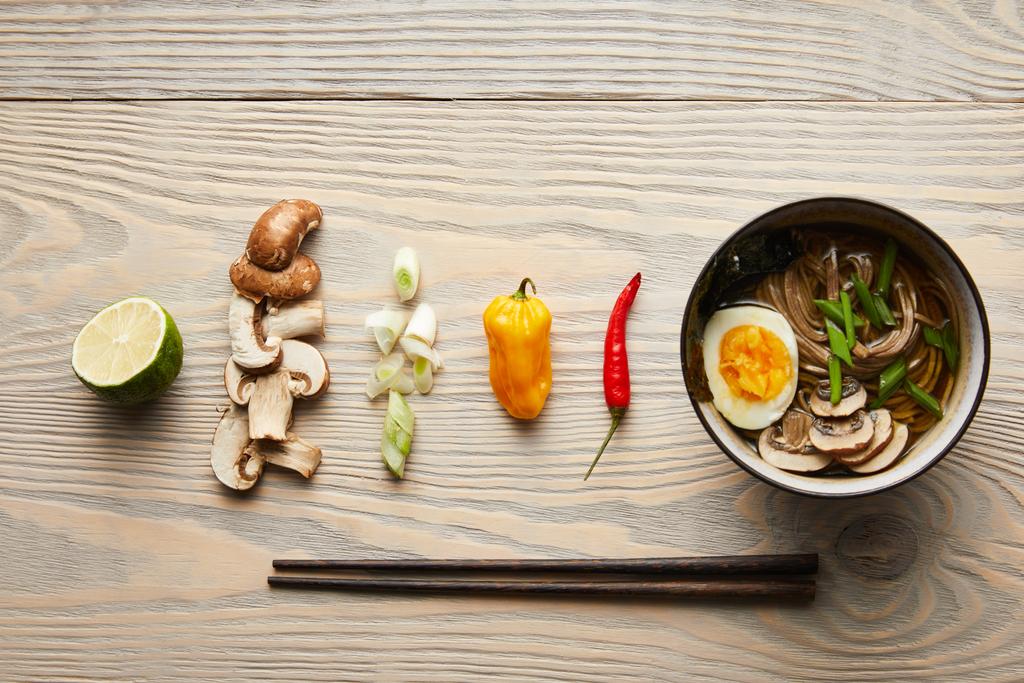 επίπεδο θέσει με παραδοσιακά πικάντικα ράμεν σε μπολ κοντά chopsticks και τα συστατικά σε ξύλινο τραπέζι - Φωτογραφία, εικόνα
