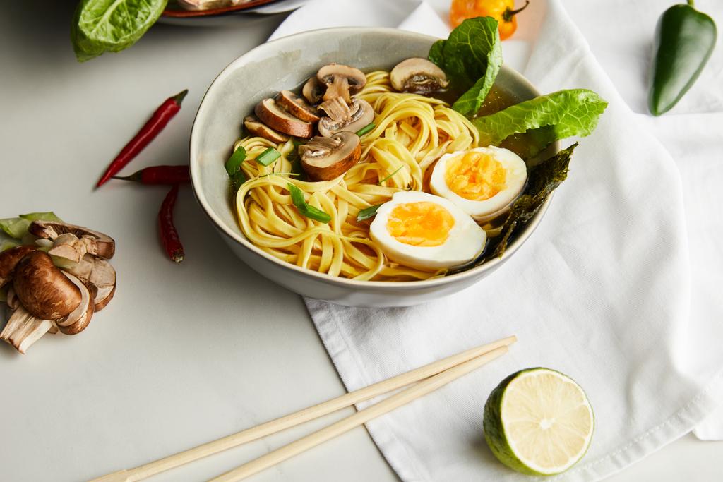 традиционный азиатский рамен в миске возле палочек для еды, салфеток и овощей на серой поверхности
 - Фото, изображение