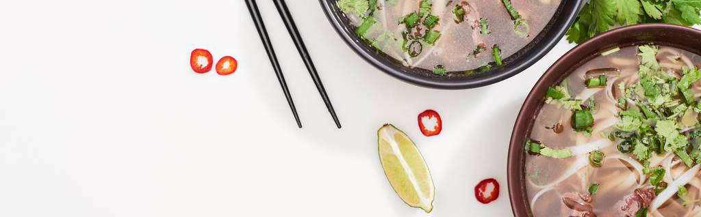 top view of pho in bowl near chopsticks, lime, chili and soy sauces and coriander on white background, panoramic shot - 写真・画像