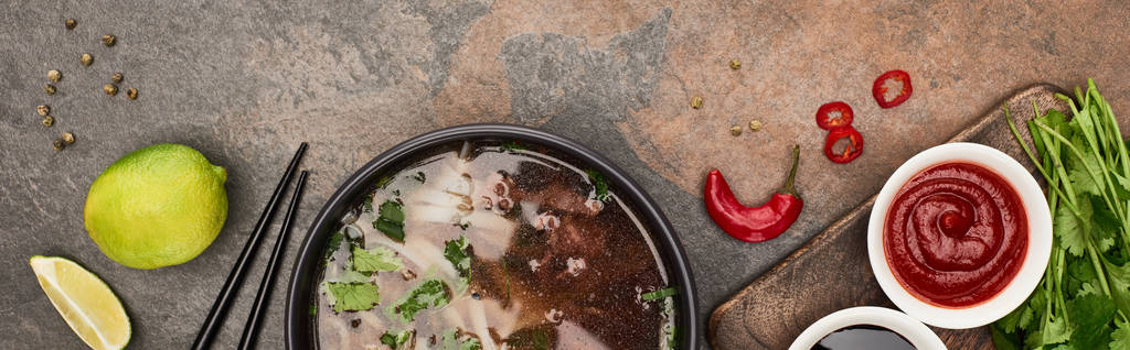 top view of pho in bowl near chopsticks, lime, chili and soy sauces and coriander on stone background, panoramic shot - Photo, Image
