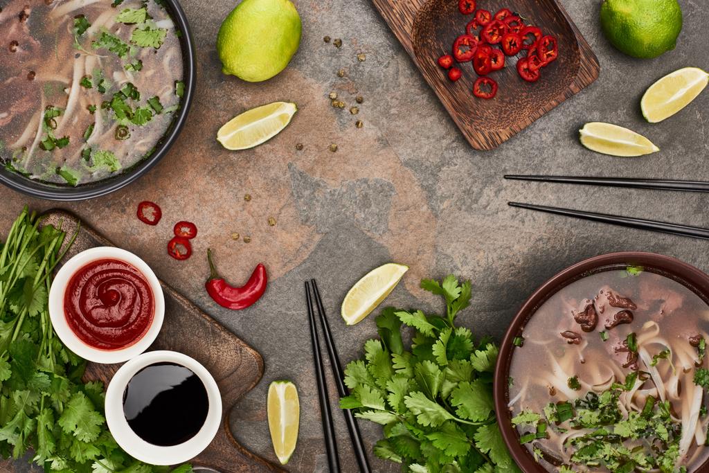 top view of pho in bowls near chopsticks, lime, chili and soy sauces and coriander on stone background - Photo, Image