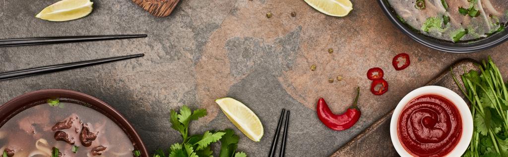 top view of pho in bowls near chopsticks, lime, chili and soy sauces and coriander on stone background, panoramic shot - Photo, Image