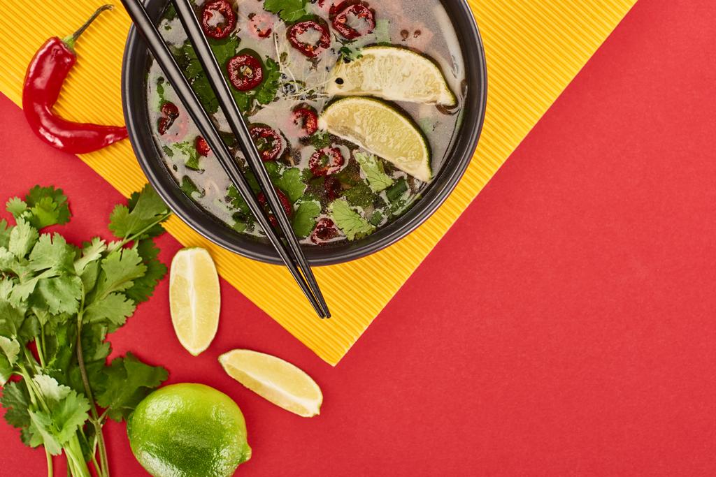 top view of pho in bowl with chopsticks near lime, chili and coriander on red and yellow background - 写真・画像