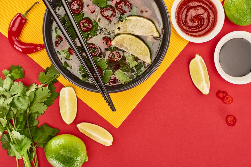 bovenaanzicht van pho in kom met eetstokjes bij sauzen, limoen, chili en koriander op rode en gele achtergrond - Foto, afbeelding