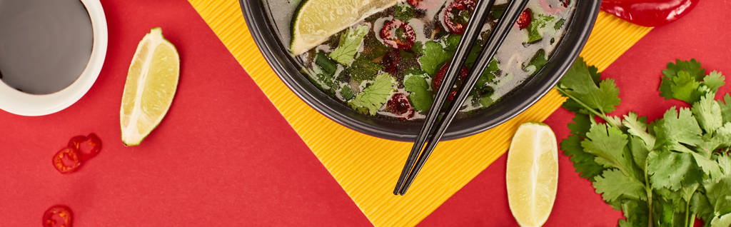 top view of pho in bowl with chopsticks near soy sauce, lime, chili and coriander on red and yellow background, panoramic shot - 写真・画像