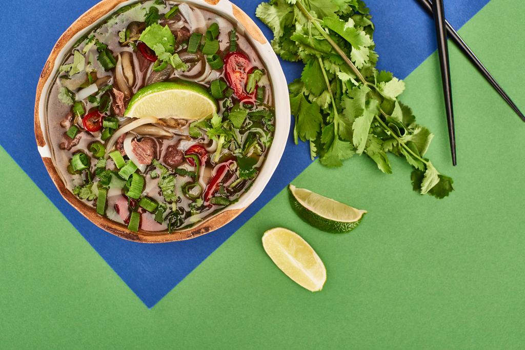 верхний вид фо в миске рядом палочки для еды, лайм, кориандр на синем и зеленом фоне
 - Фото, изображение