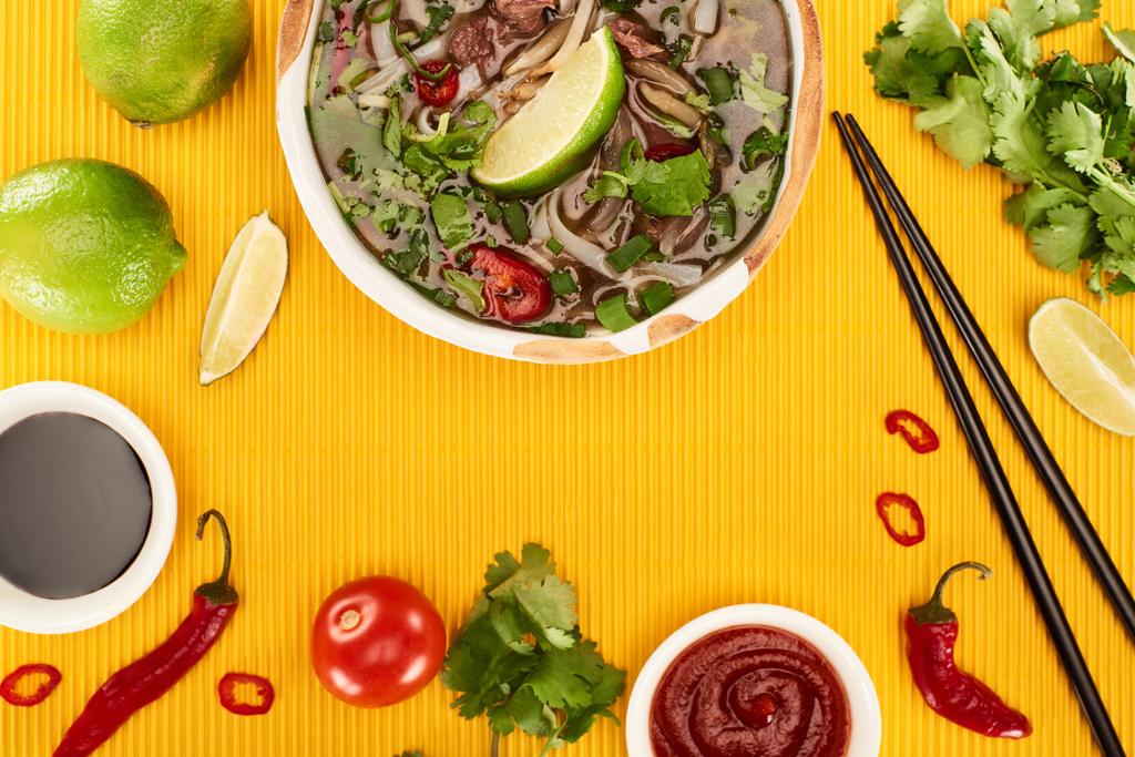 top view of pho in bowl near chopsticks, lime, chili and soy sauces and coriander on yellow textured background - Photo, Image