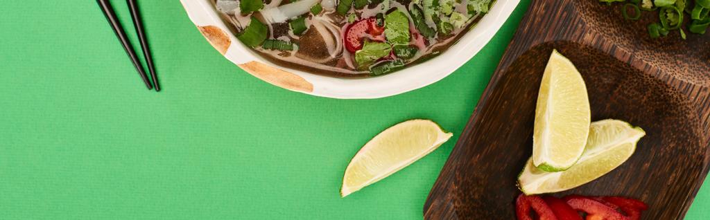 top view of pho in bowl near chopsticks and wooden board with lime on green background, panoramic shot - Photo, Image