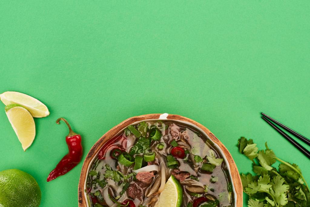 top view of pho in bowl near chopsticks, lime, chili and coriander on green background - 写真・画像