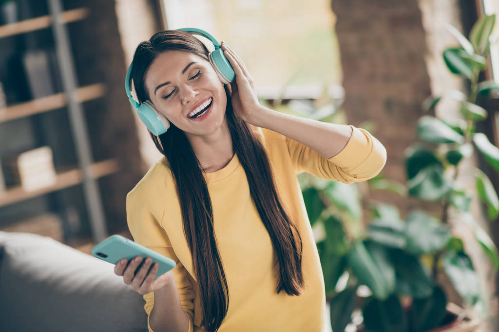 Portret pozytywnej wesołej dziewczyny słuchać muzyki na jej smartfonie wybrać melodię cieszyć się piosenką nosić żółty sweter w pokoju apartament - Zdjęcie, obraz