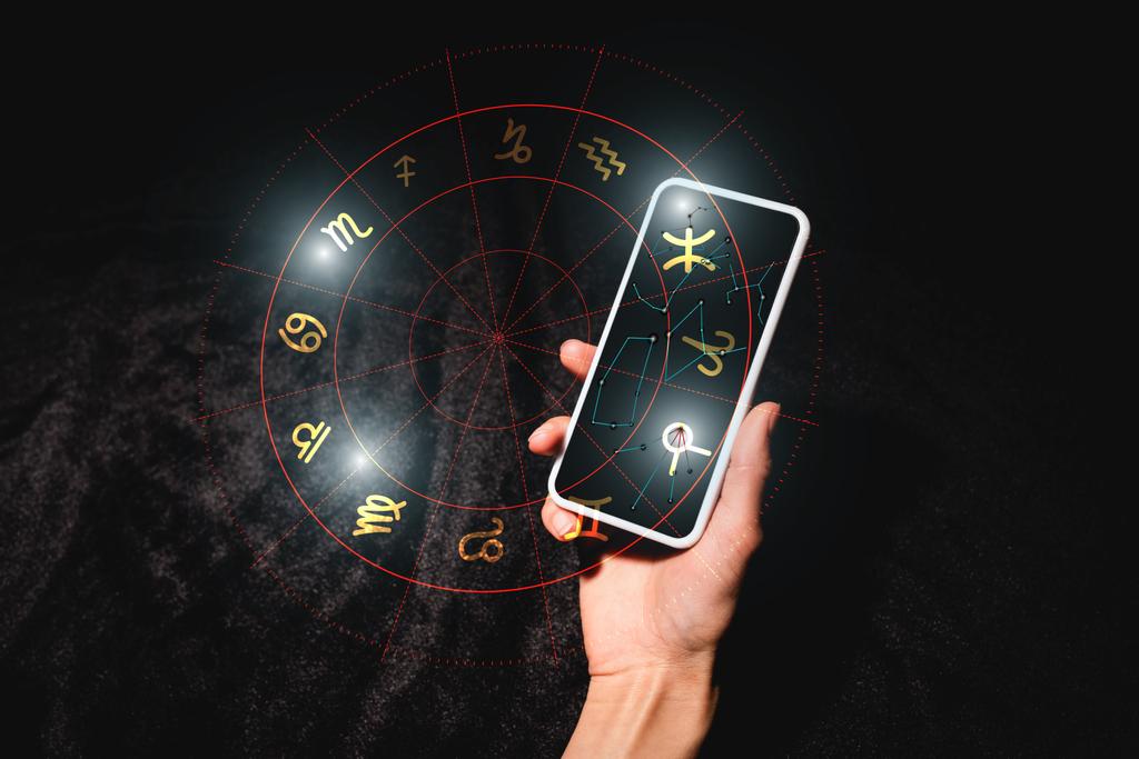 黒いベルベットの布の上に星座のついたスマートフォンを持っている占星術師のクロップドビュー - 写真・画像