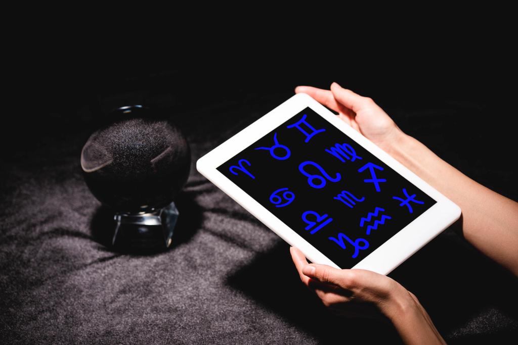 обрезанный вид астролога, держащего цифровой планшет с знаками зодиака возле хрустального шара на черной бархатной ткани
 - Фото, изображение