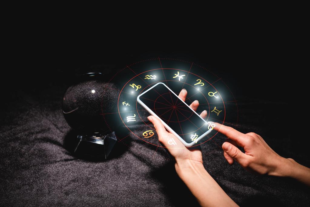 обрізаний вид астролога, що тримає смартфон із знаками зодіаку біля кришталевого м'яча на чорній оксамитовій тканині
 - Фото, зображення