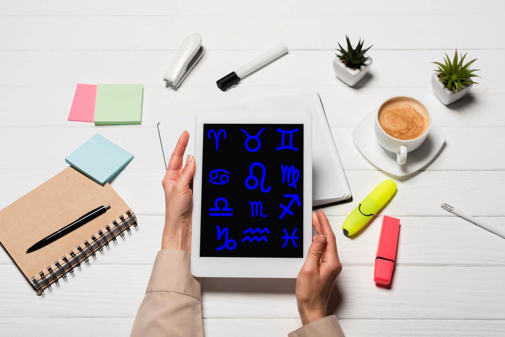 vista recortada de la mujer sosteniendo tableta digital con signos del zodiaco en el lugar de trabajo con suministros de oficina y café
 - Foto, imagen