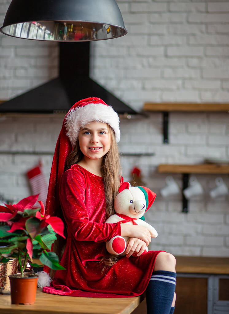 Счастливого Рождества. Милая маленькая девочка в шляпе Санты держит плюшевого мишку, сидящего на кухне, ожидая праздника
. - Фото, изображение