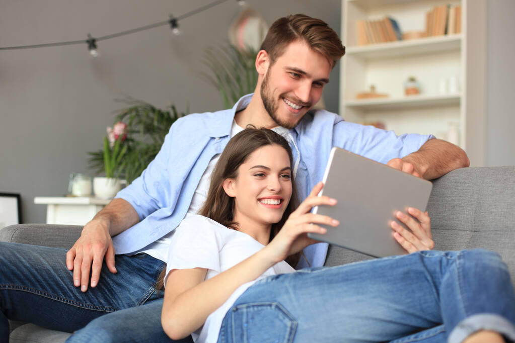 Junges Paar betrachtet Medieninhalte online auf einem Tablet sitzend auf einem Sofa im Wohnzimmer. - Foto, Bild