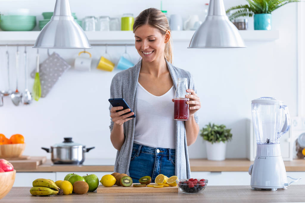 Hezká mladá žena pomocí svého mobilního telefonu při pití ovocné šťávy v kuchyni doma. - Fotografie, Obrázek