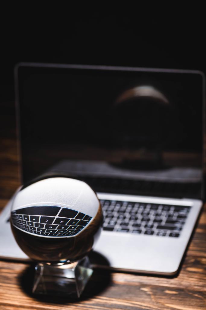 selective focus of crystal ball near laptop on wooden table on black background - Photo, Image