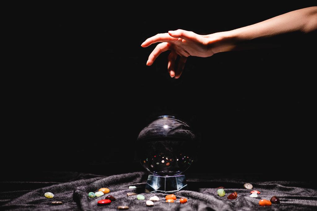 cropped άποψη του χεριού μάντισσα πάνω από κρυστάλλινη σφαίρα με πέτρες telling τύχη σε μαύρο βελούδο ύφασμα απομονώνονται σε μαύρο - Φωτογραφία, εικόνα