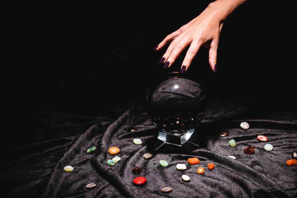 vista ritagliata della mano indovino sopra sfera di cristallo con pietre predittive di fortuna su panno di velluto nero isolato su nero
 - Foto, immagini