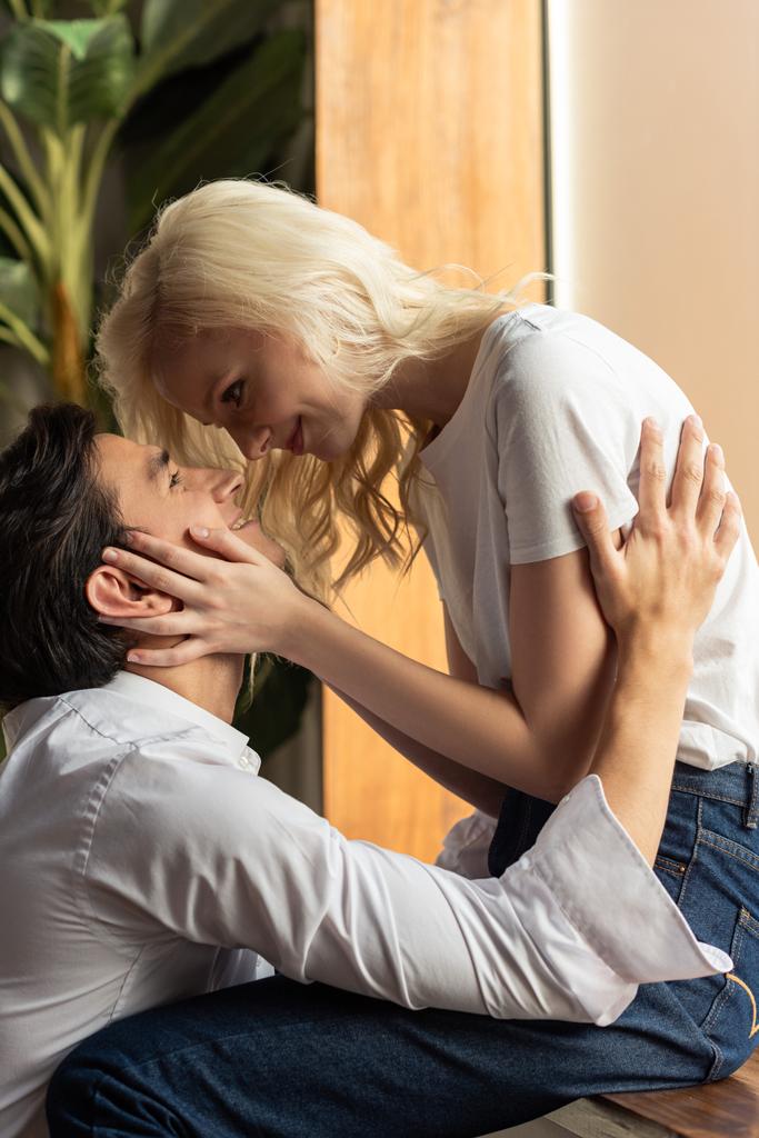 Side view of smiling blonde girl touching boyfriend while sitting on windowsill at home - Photo, Image