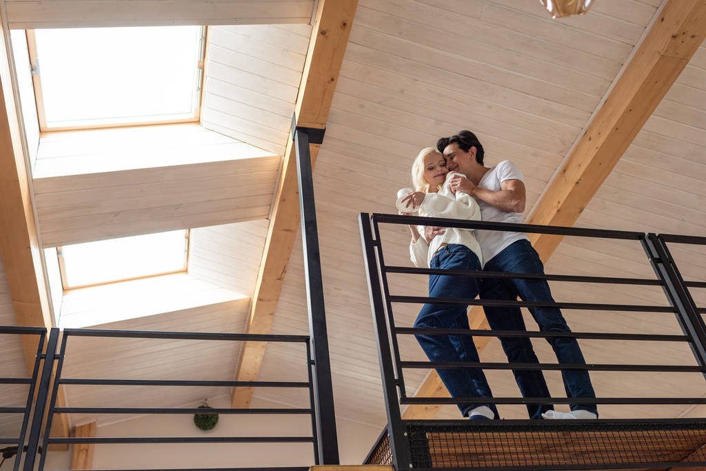 Χαμηλή γωνία άποψη του νεαρού ζευγαριού αγκαλιάζει στο μπαλκόνι και το κορίτσι κρατώντας φλιτζάνι καφέ - Φωτογραφία, εικόνα