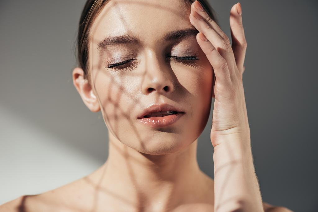 πορτρέτο της τρυφερής γυμνής γυναίκας με κλειστά μάτια και σκιές στο πρόσωπο σε γκρι - Φωτογραφία, εικόνα