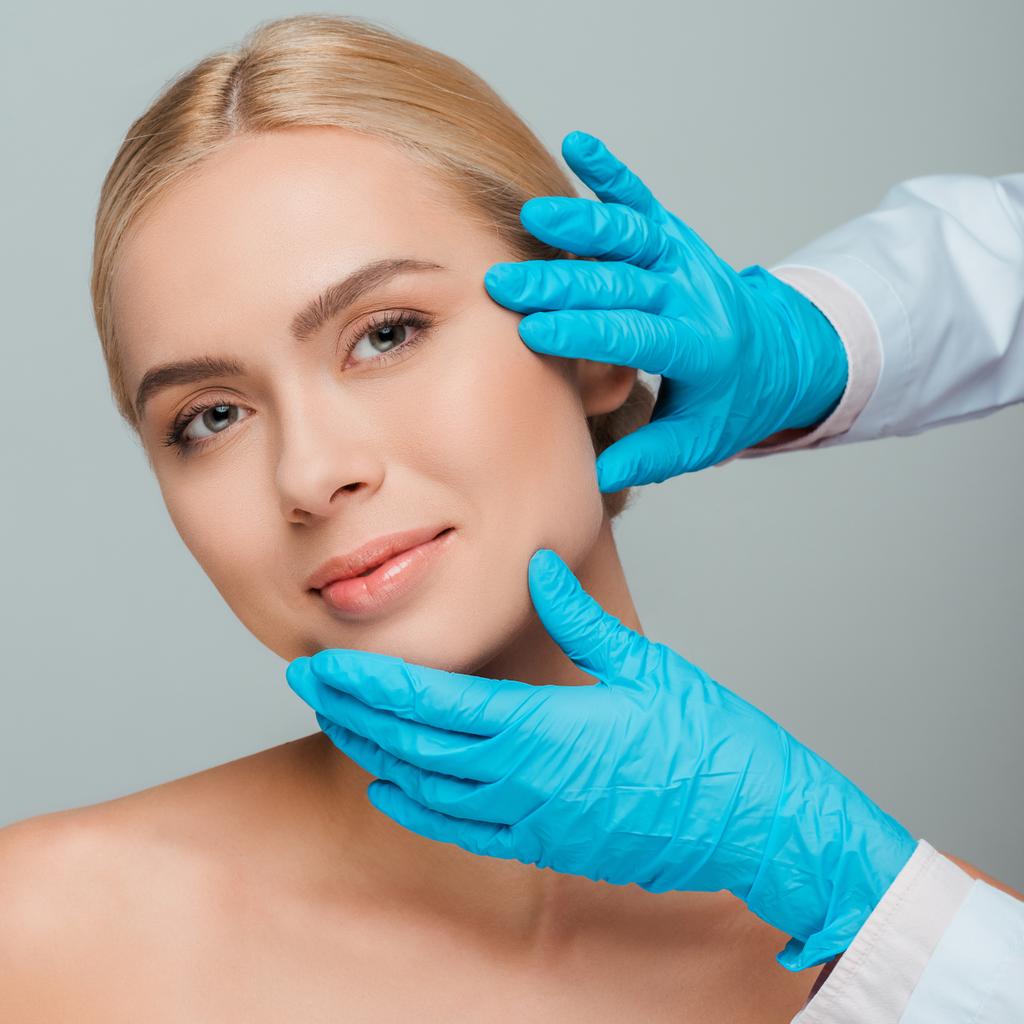 abgeschnittene Ansicht der Kosmetikerin in Latex-Handschuhen berührt Gesicht der jungen Frau isoliert auf grau  - Foto, Bild