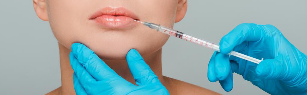 Panoramaaufnahme einer Kosmetikerin in Latexhandschuhen, die eine Spritze in der Nähe der Lippen einer Frau hält, isoliert auf grau  - Foto, Bild