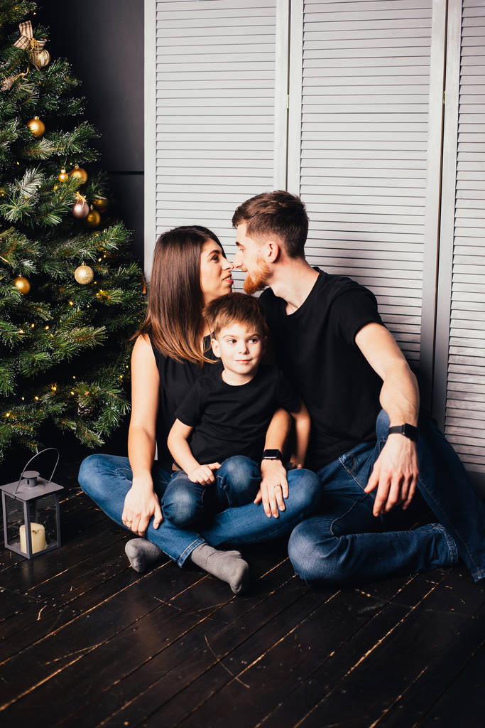 счастливая молодая семья с одним ребенком, сидящая вместе у новогодней елки
 - Фото, изображение