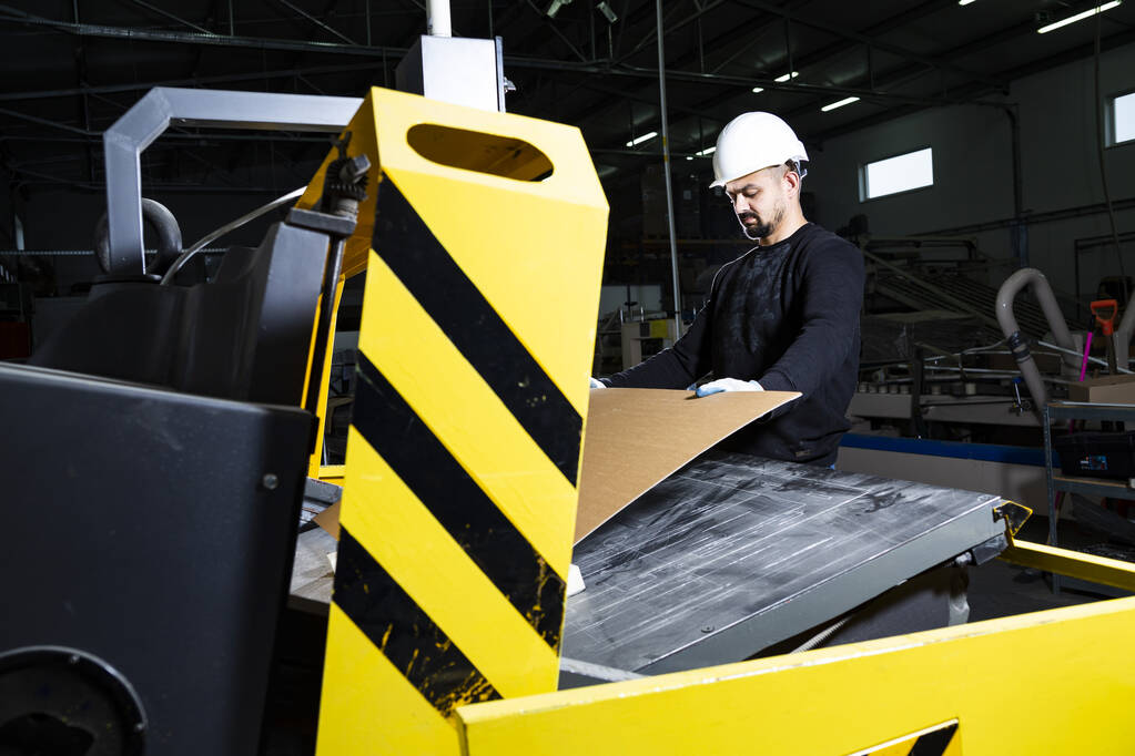 Παραγωγή χαρτοκιβωτίων. Εργαζόμενος κόβει χαρτοκιβώτιο με ένα μηχάνημα διάτρησης. Κοπτικό μηχάνημα χαρτιού.  - Φωτογραφία, εικόνα