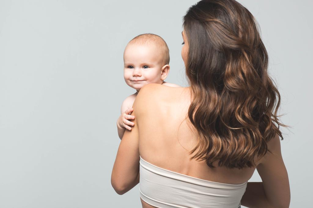 vista trasera de la joven madre sosteniendo al bebé niño, aislado en gris
 - Foto, Imagen