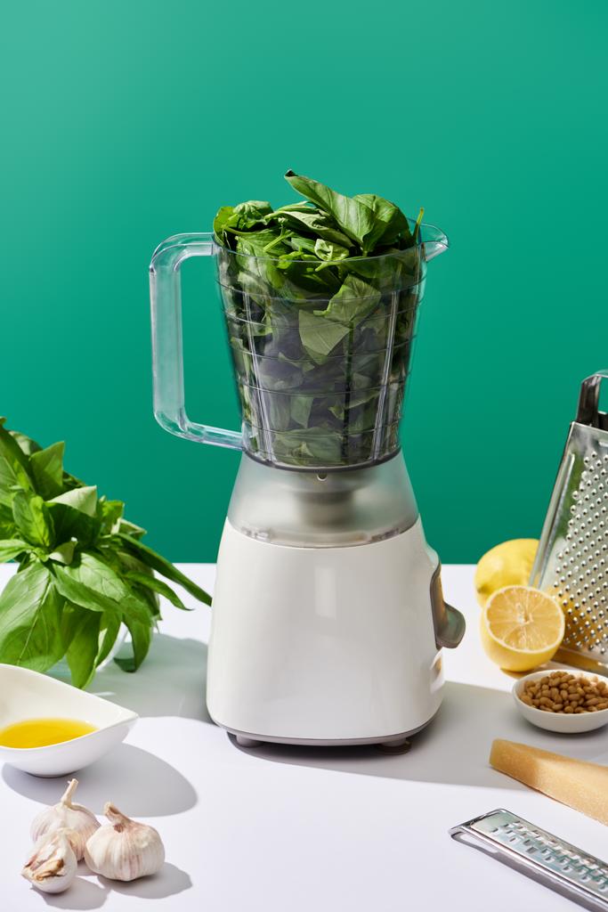 pesto saus grondstoffen en keukenmachine met basilicum bladeren op witte tafel geïsoleerd op groen - Foto, afbeelding