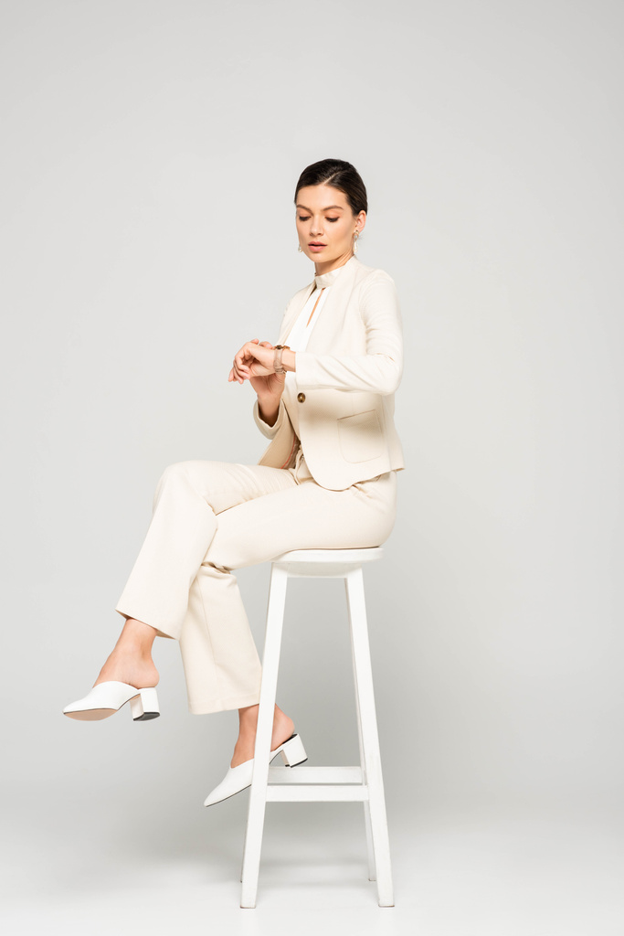 elegante zakenvrouw in wit pak kijken naar horloge terwijl zitten op kruk, op grijs - Foto, afbeelding