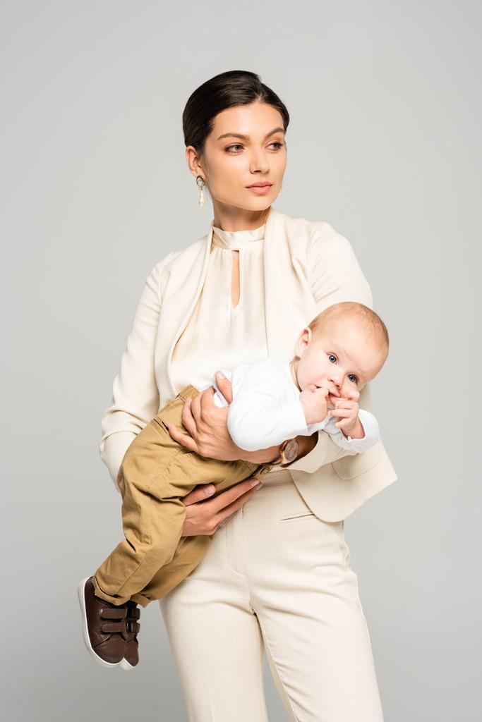 schöne Geschäftsfrau mit entzückendem Baby an den Händen, isoliert auf grau - Foto, Bild