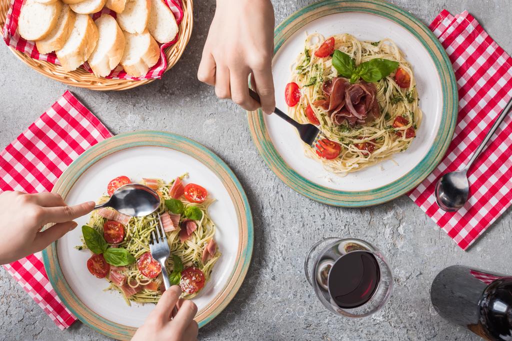 levágott kilátás a nők eszik Pappardelle paradicsommal, bazsalikom és prosciutto közelében baguette, vörösbor szürke felületen - Fotó, kép