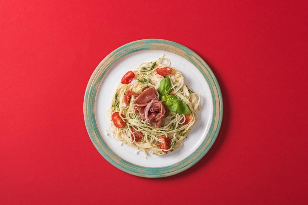 Draufsicht auf gekochte Pappardelle mit Tomaten, Basilikum und Prosciutto auf Teller auf rotem Hintergrund - Foto, Bild